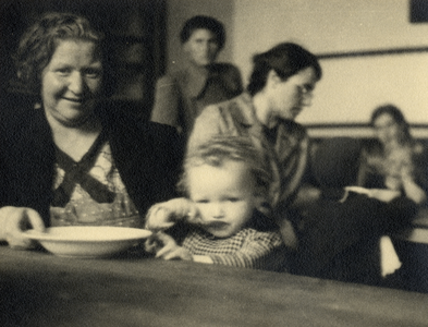 811775 Afbeelding van een moeder met kind aan een, uit een gaarkeuken afkomstige, gezamelijke maaltijd in een lokaal in ...